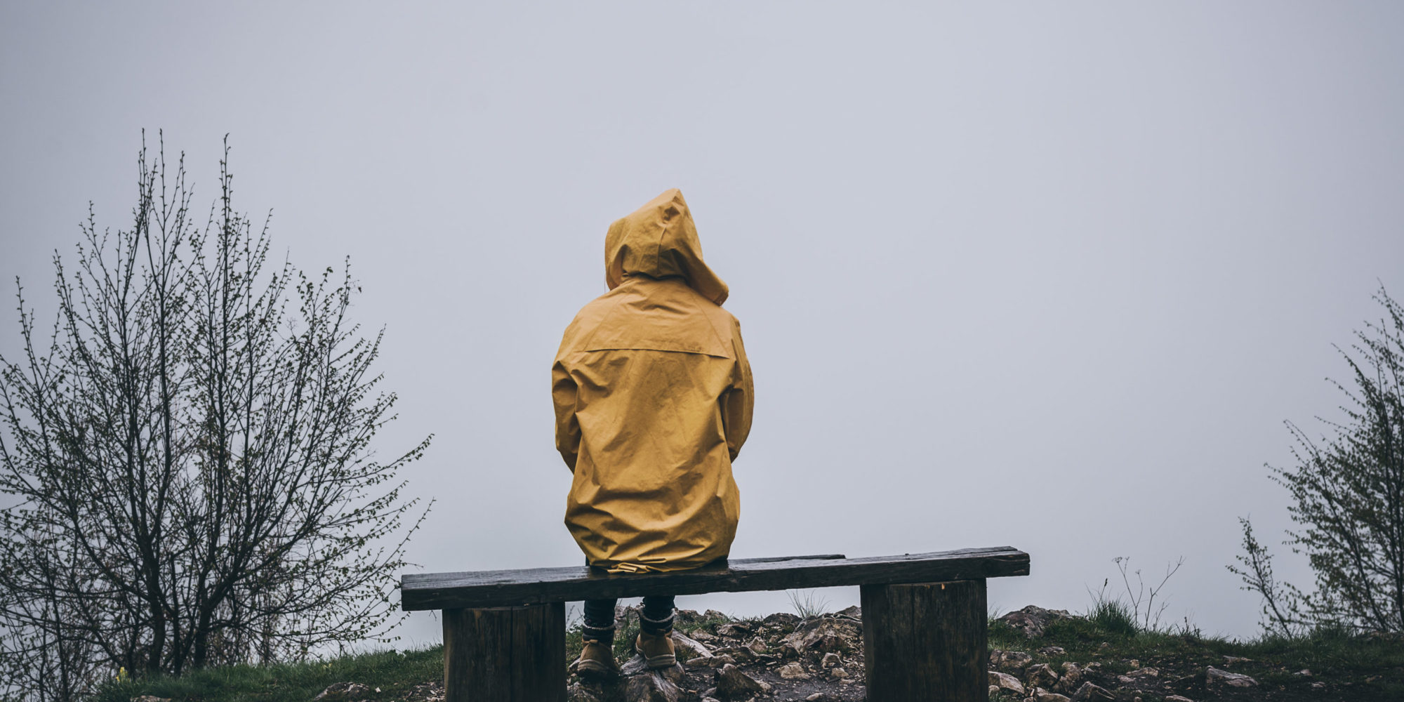 Person sitzt mit gelber Regenjacke auf einer Holzbank. Überall Nebel.